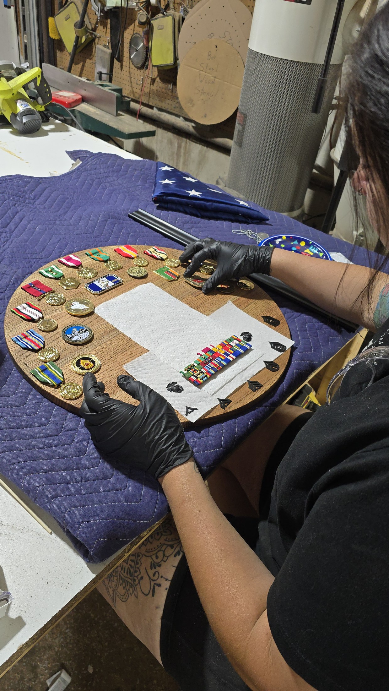 Gloved hands arrange medals and insignia around a folded American flag on a circular wood display in the workshop.