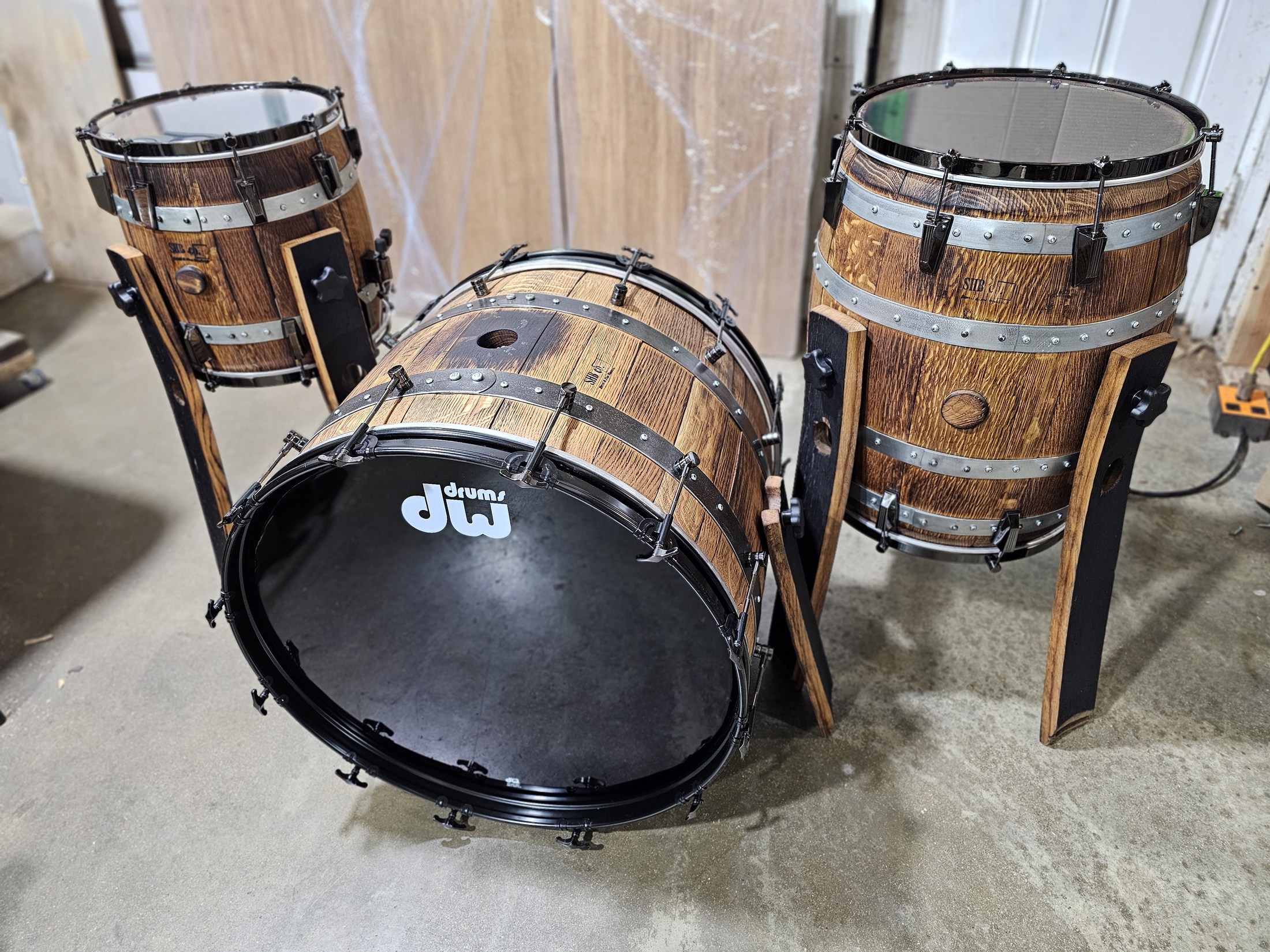Three-piece drum kit made from wooden barrels, with black drumheads, metal hoops and tall wood supports.