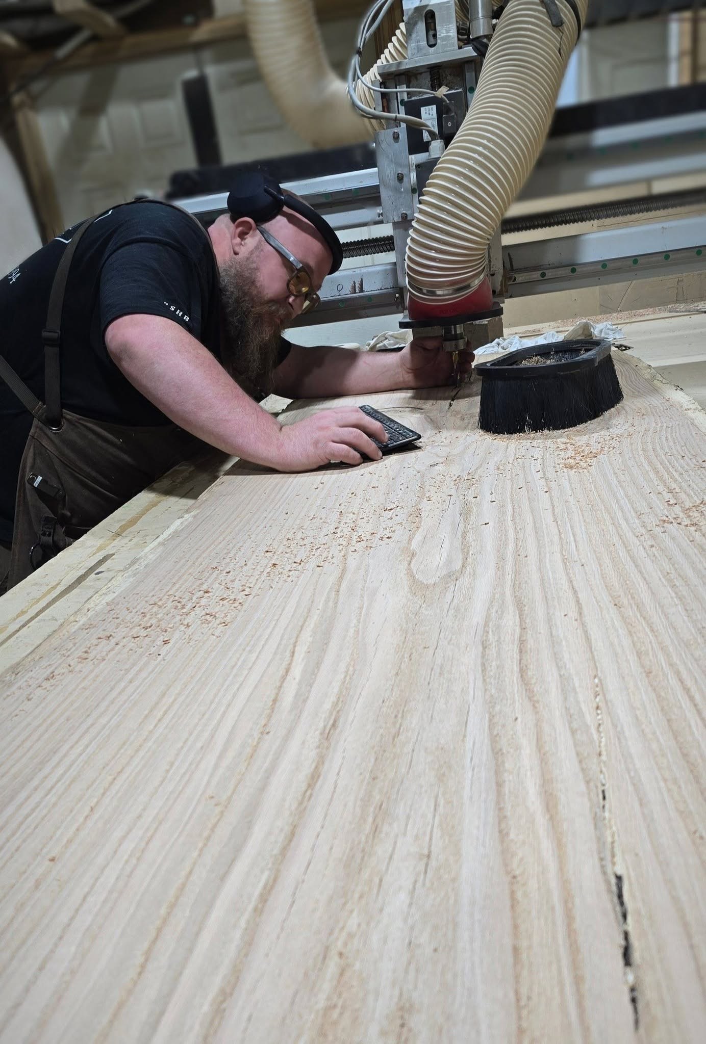 A woodworker checks a broad black-oak slab beneath the CNC spindle and dust-extraction hose in the SHB workshop.