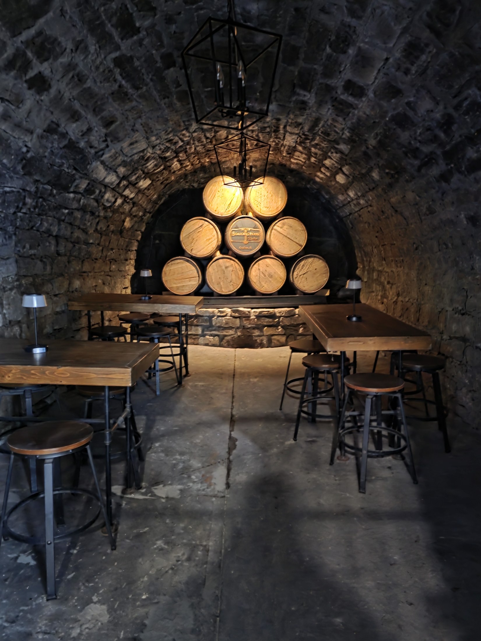 Barrel pyramid with a central Boarding House Bistro cap at the end of a vaulted stone cellar with tables and stools.