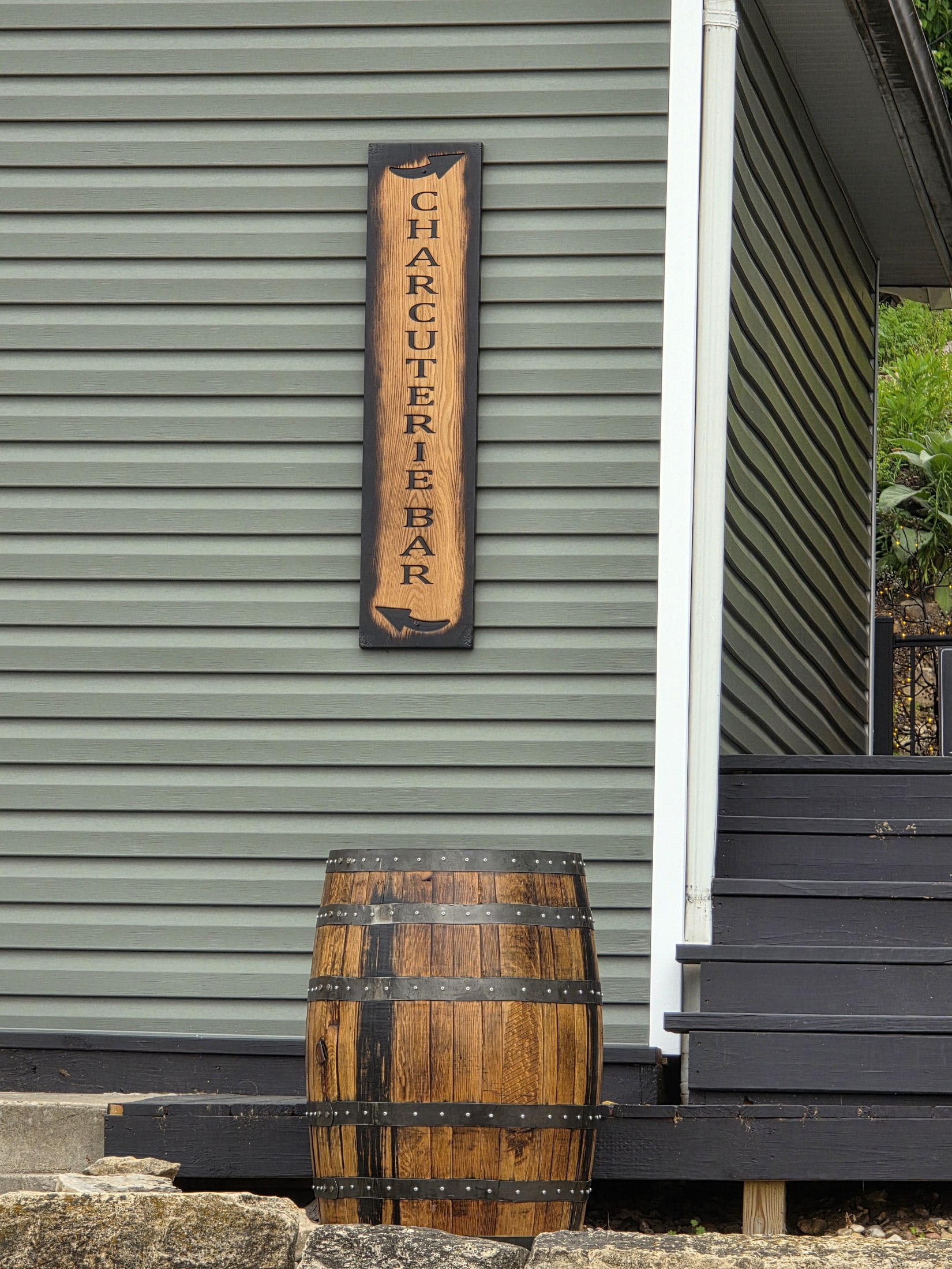Vertical wooden Charcuterie Bar sign above a barrel, mounted on green exterior siding.