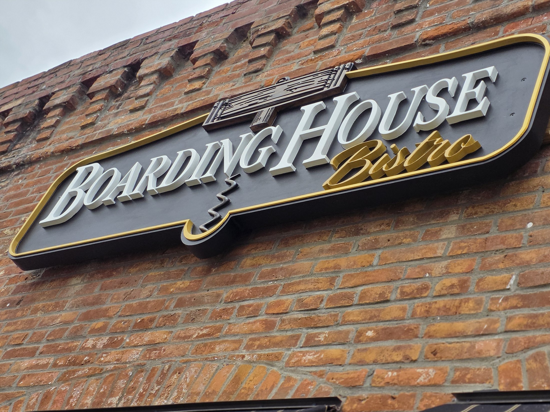 Angled view of the Boarding House Bistro sign, with raised white lettering, gold script and a corkscrew detail on the brick frontage.