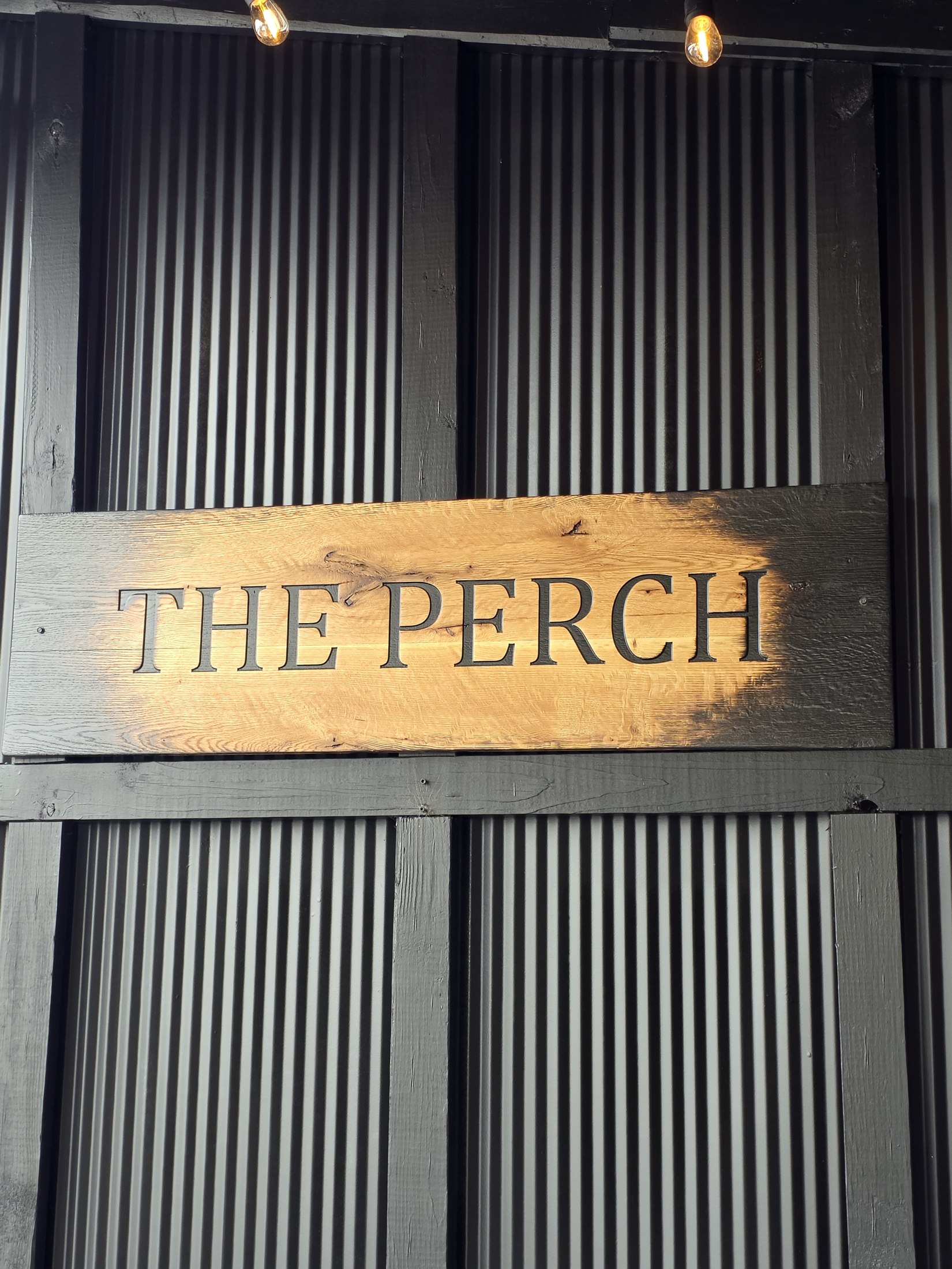 Horizontal wooden The Perch sign mounted over corrugated siding.