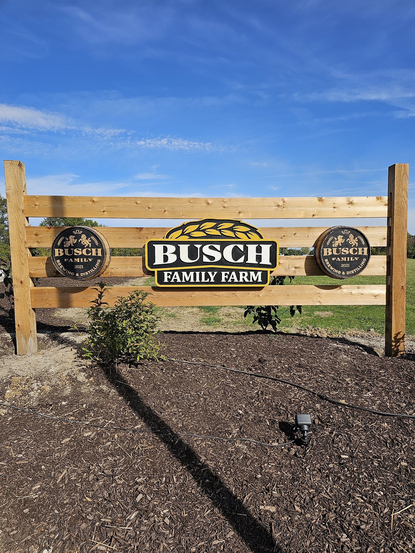Black, white and gold Busch Family Farm sign flanked by two round barrel-cap signs on a timber fence.