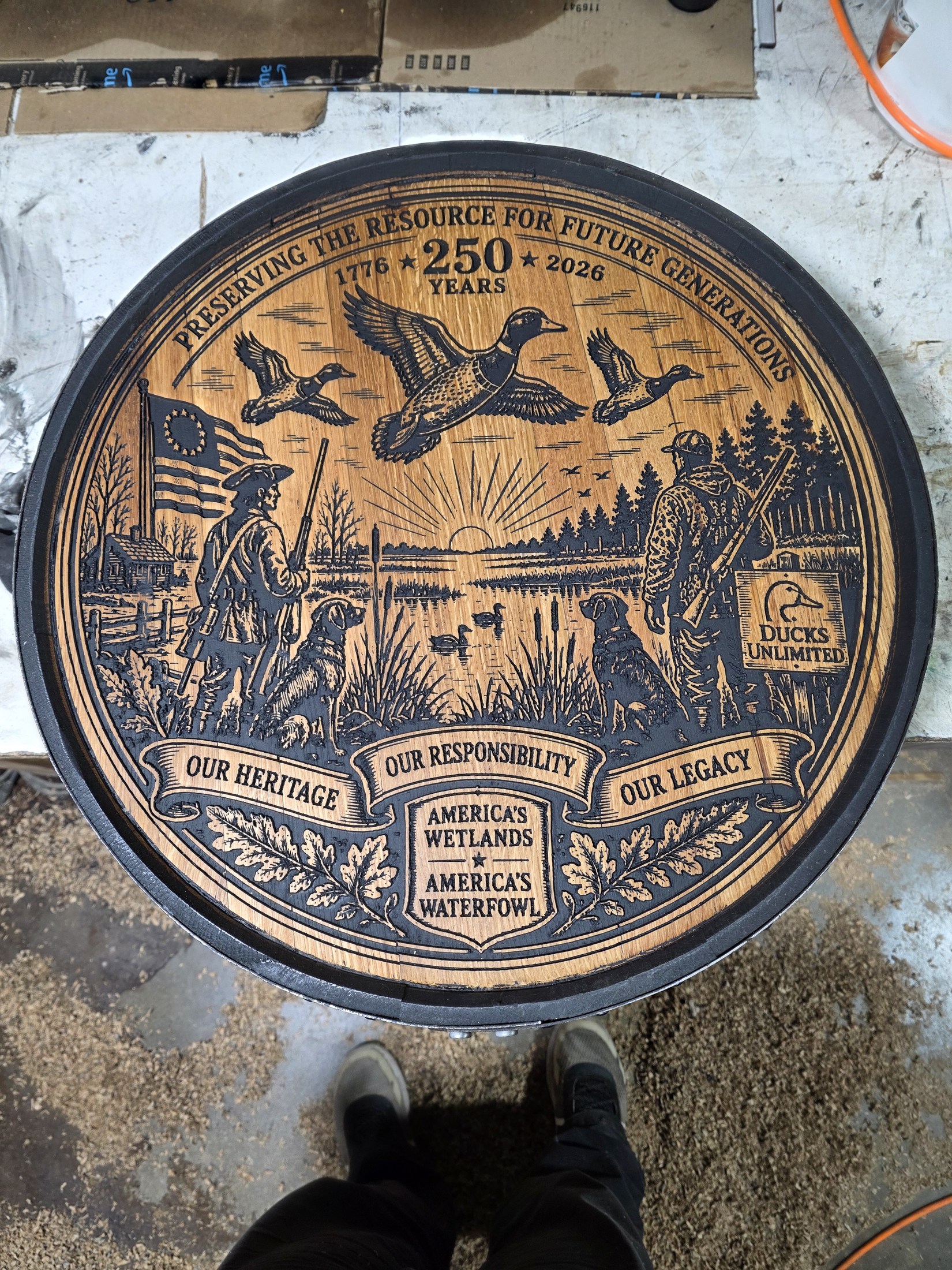 Finished circular wood carving with flying ducks, wetlands, conservation scenes and America 250 lettering, photographed in the workshop.