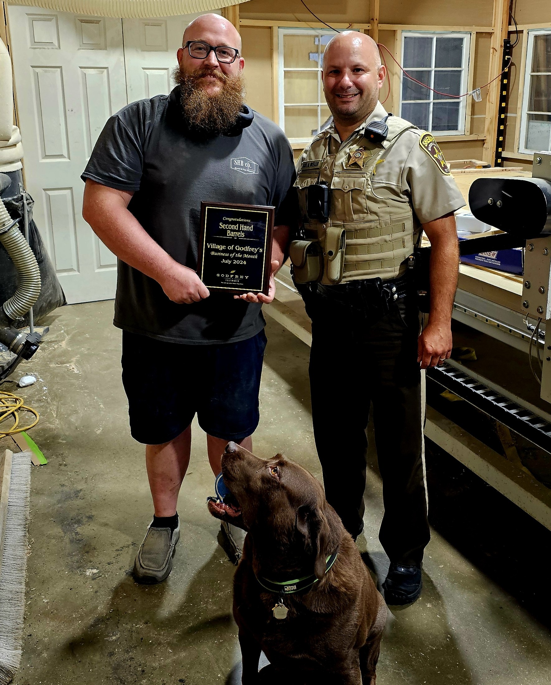 Ryan holds the Godfrey Business of the Month plaque beside Lt. Weller in the SHB shop, with the shop dog in front.