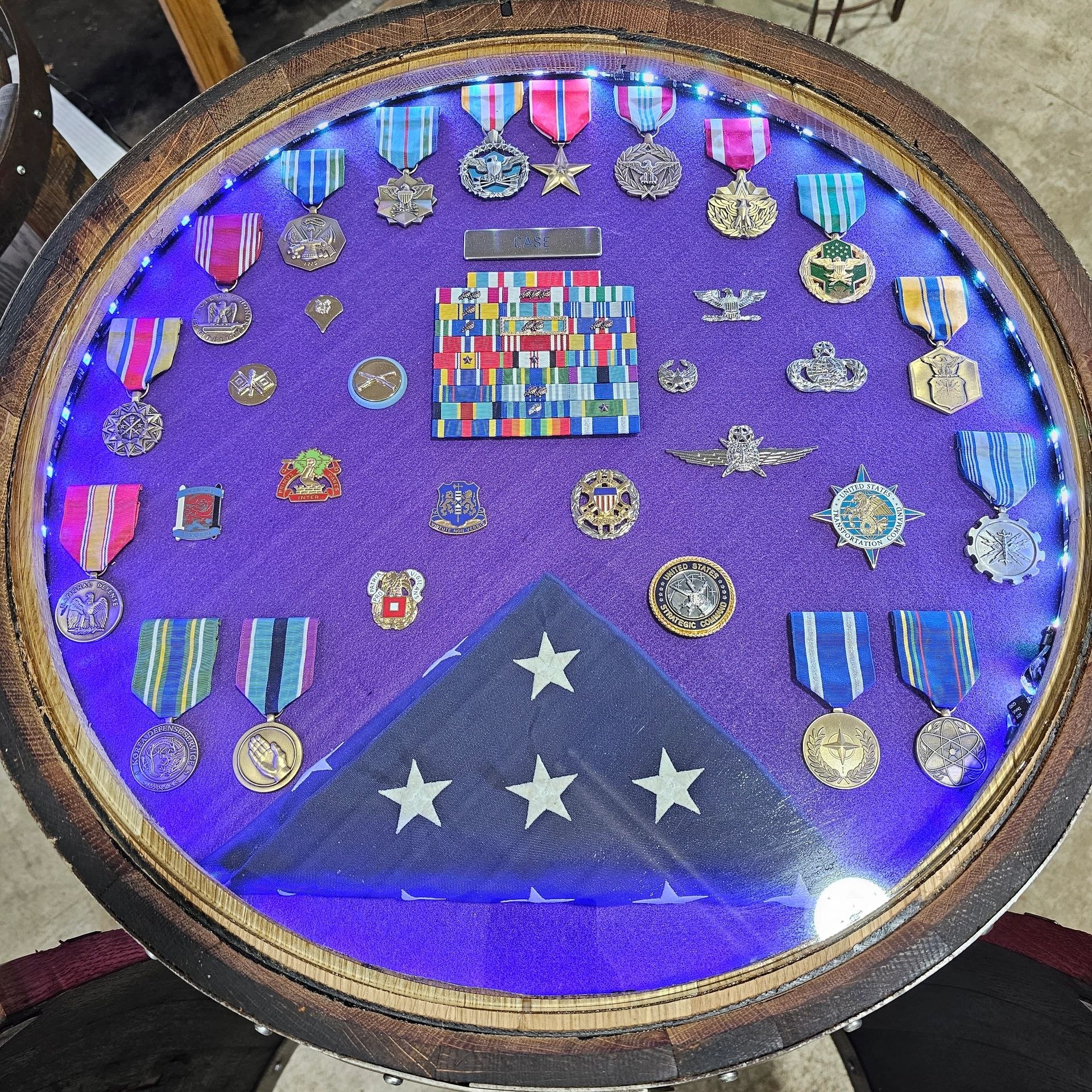 Circular barrel shadow box displaying military medals, patches and a folded flag against a blue background.