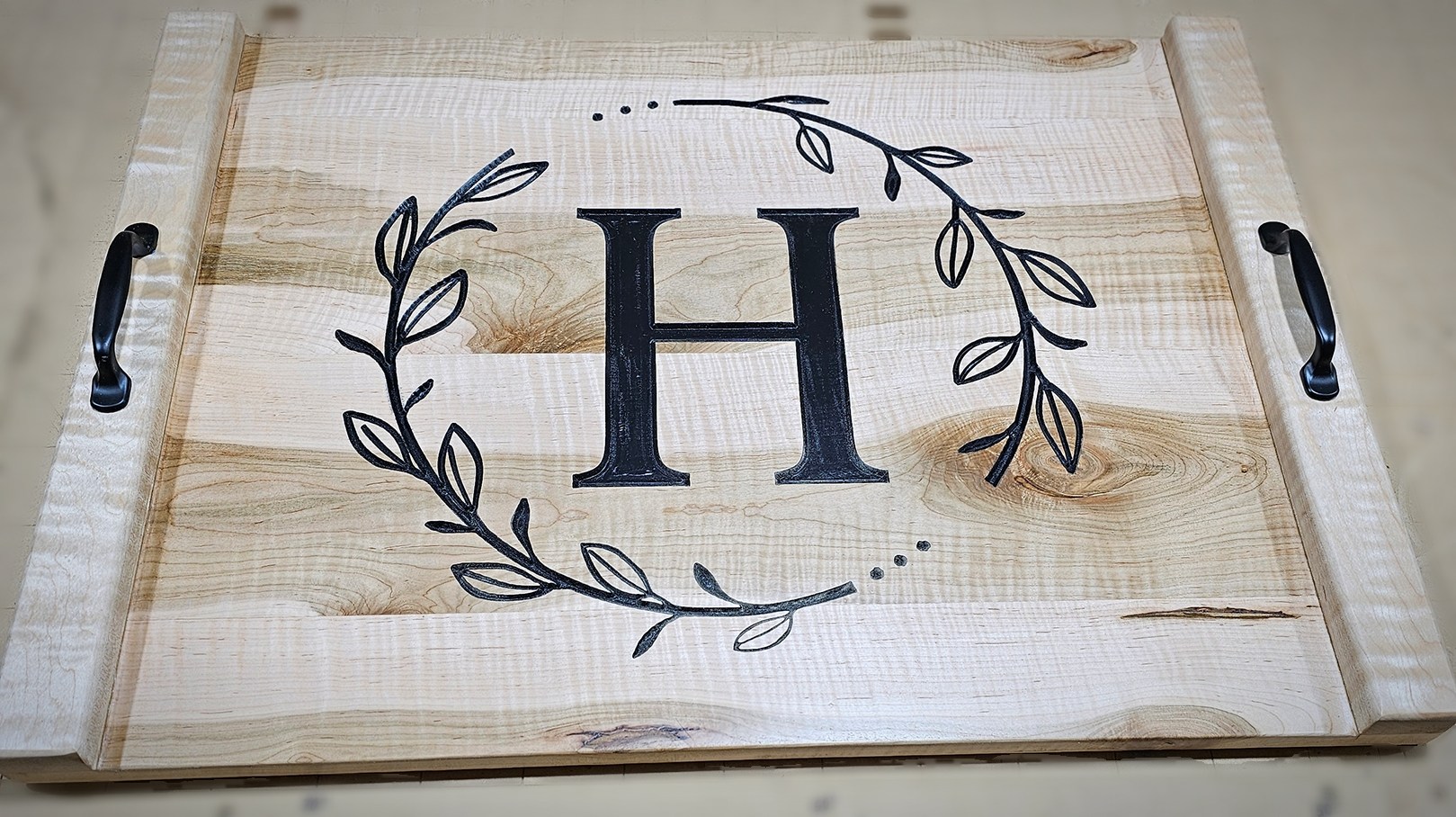 Figured ambrosia maple noodle board with black handles and an engraved H inside a leafy wreath.