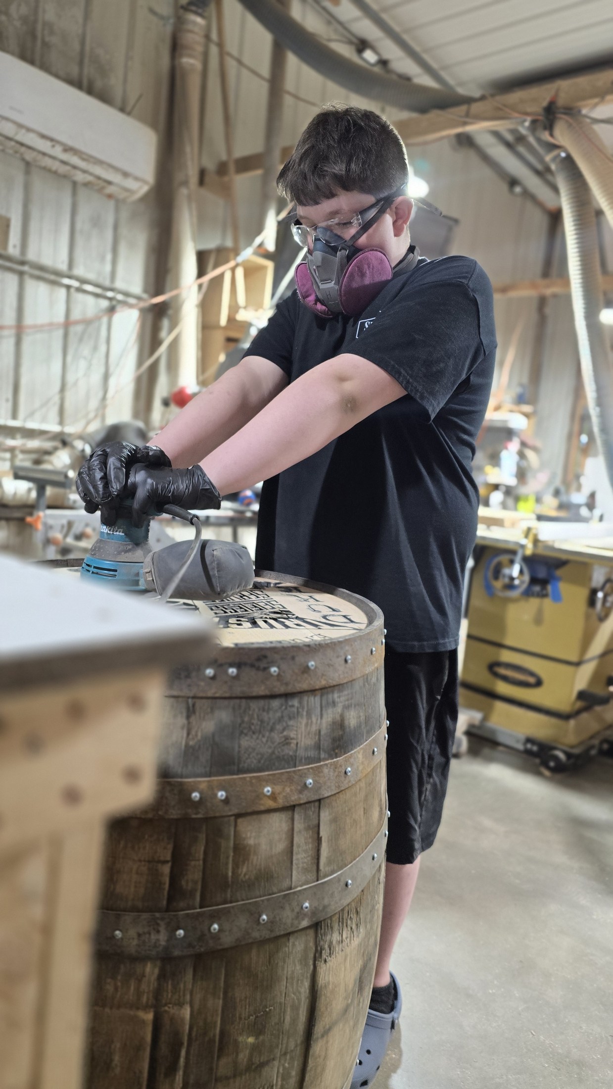 A young family member wearing a respirator, eye protection and gloves while sanding the top of a barrel in the shop.