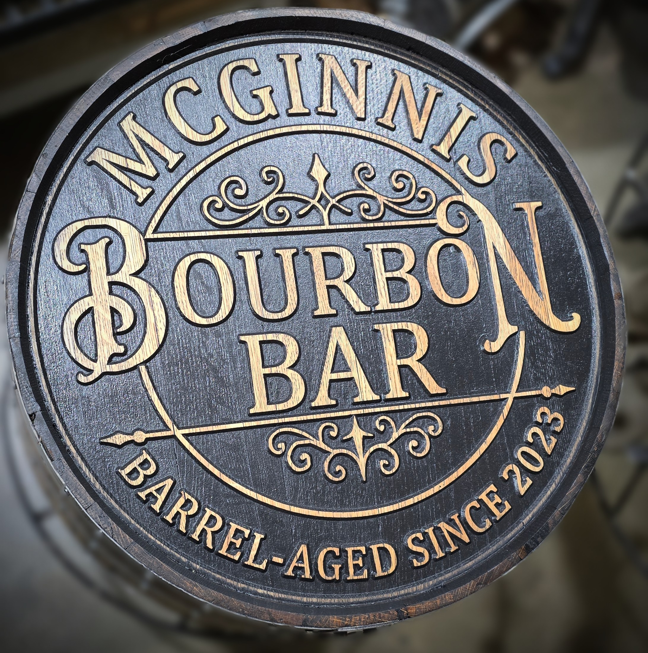 Round engraved wood cap reading McGinnis Bourbon Bar, surrounded by scrollwork and a dark circular border.