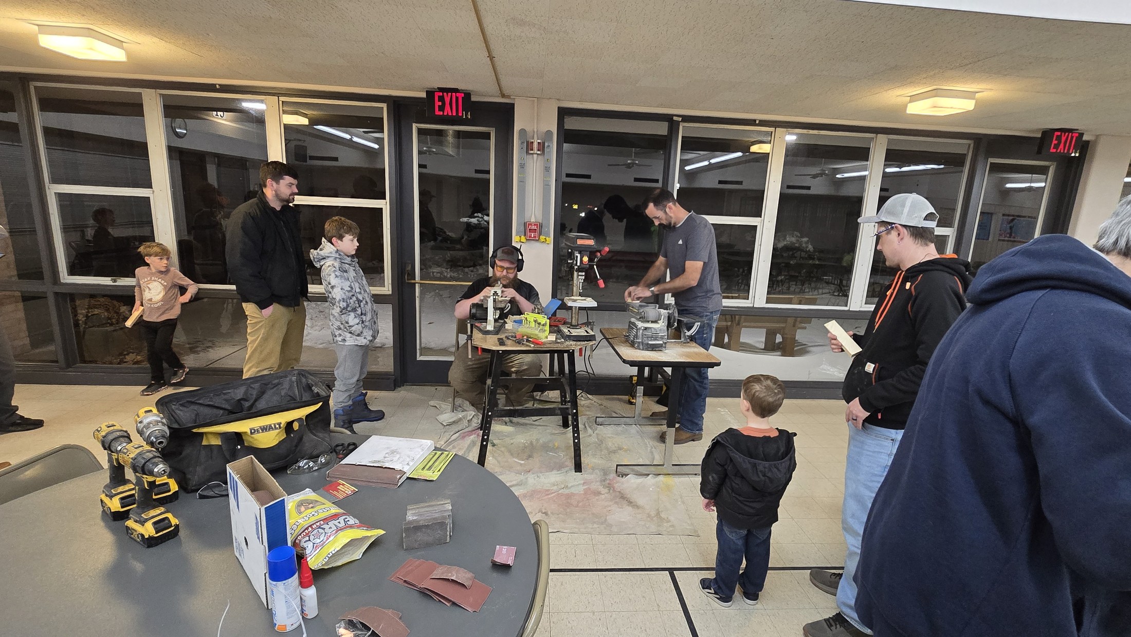 Children and adults gather around temporary woodworking stations during a local Cub Scout Pinewood Derby build night.