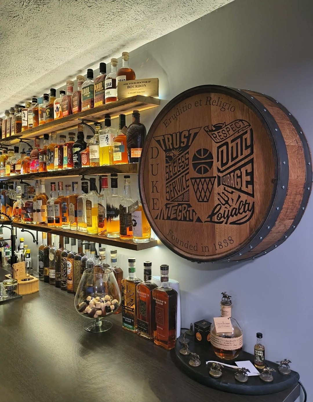Engraved Root family barrel cap beside illuminated bottle shelves and the dark counter in the home bar.