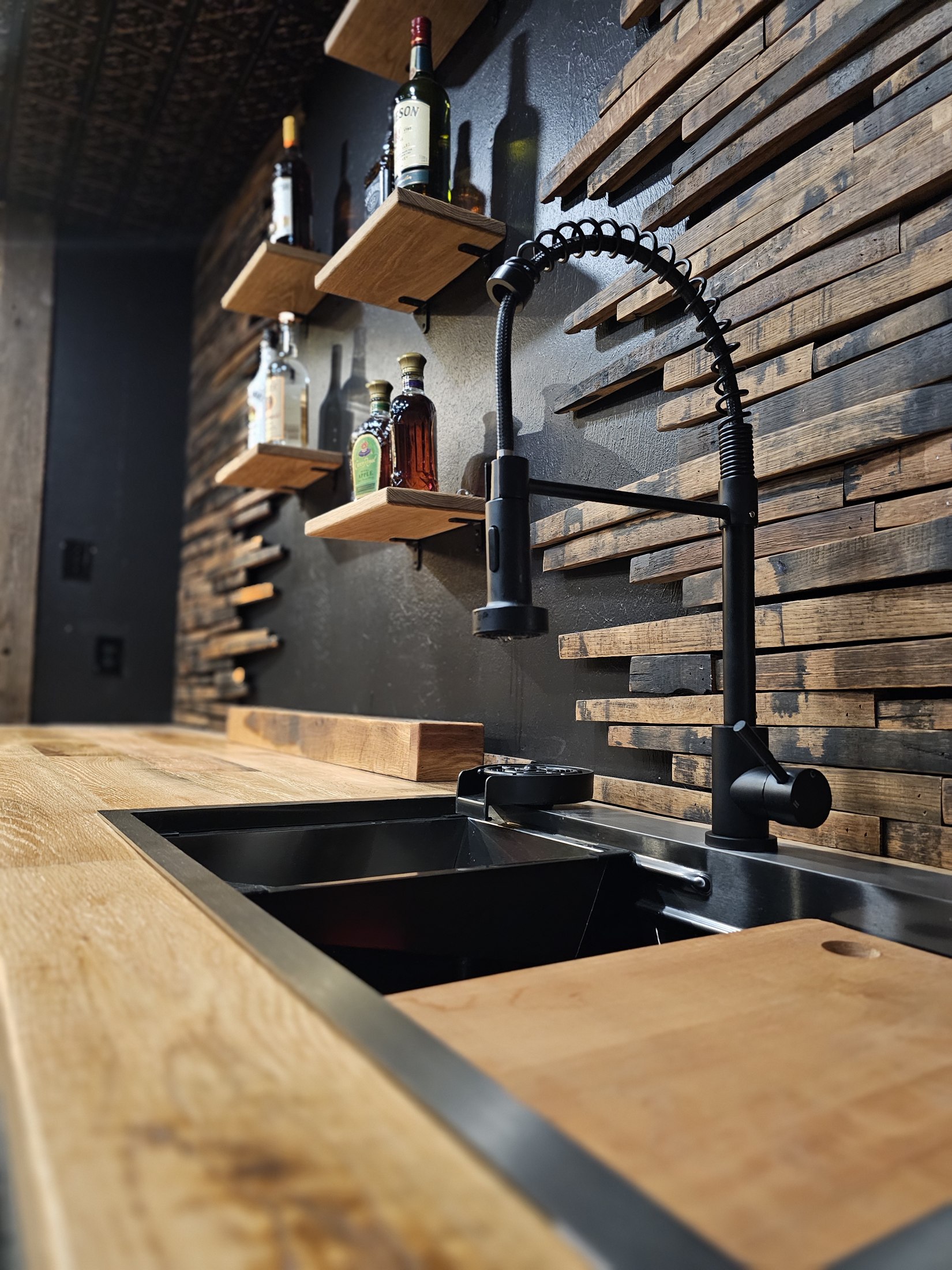 Dark sink and faucet set into a wood counter beside a stave-covered wall.