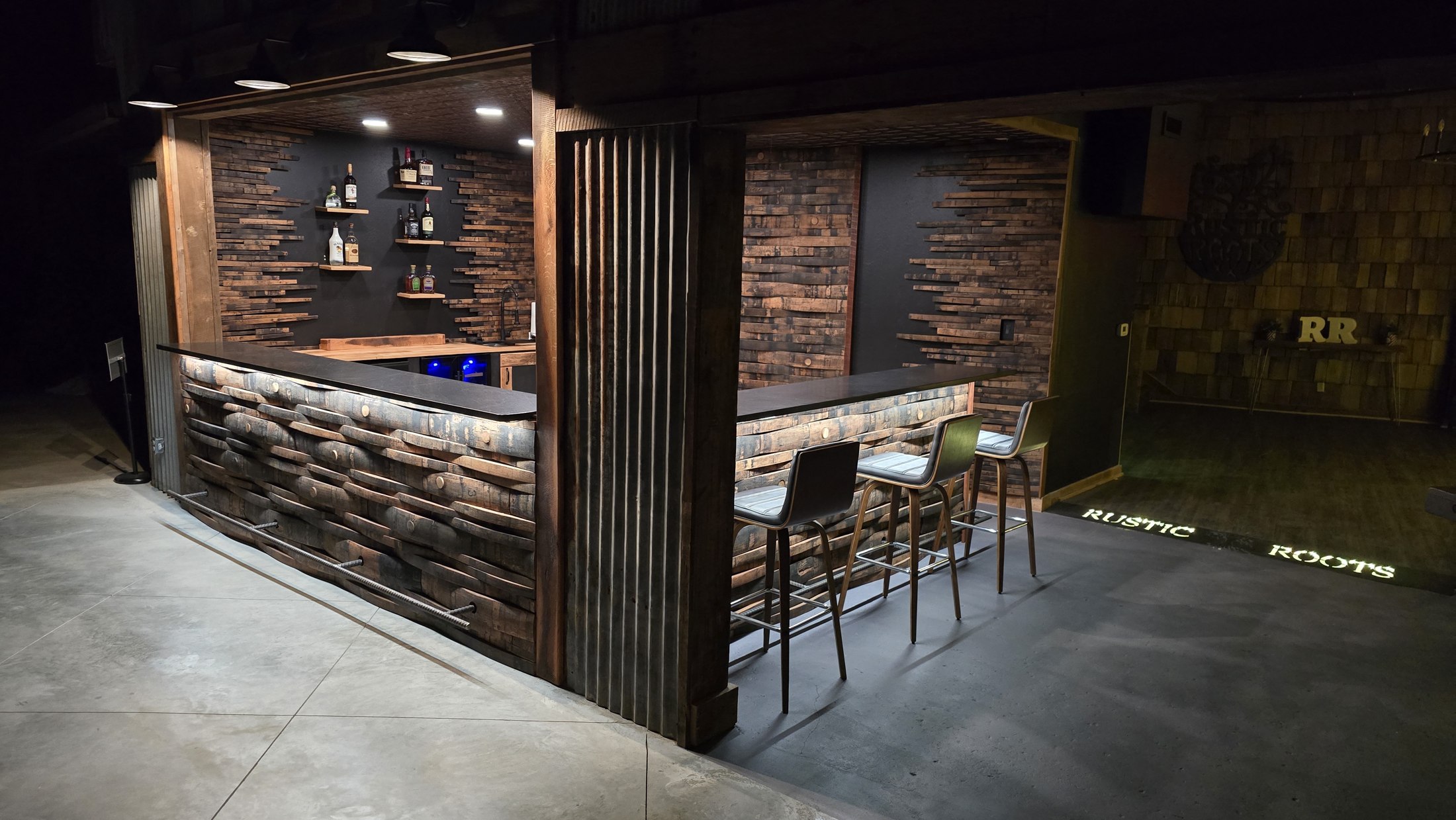 Wide corner view of the Rustic Roots bar with staggered stave fronts, dark counters, a metal foot rail and bottle shelving.