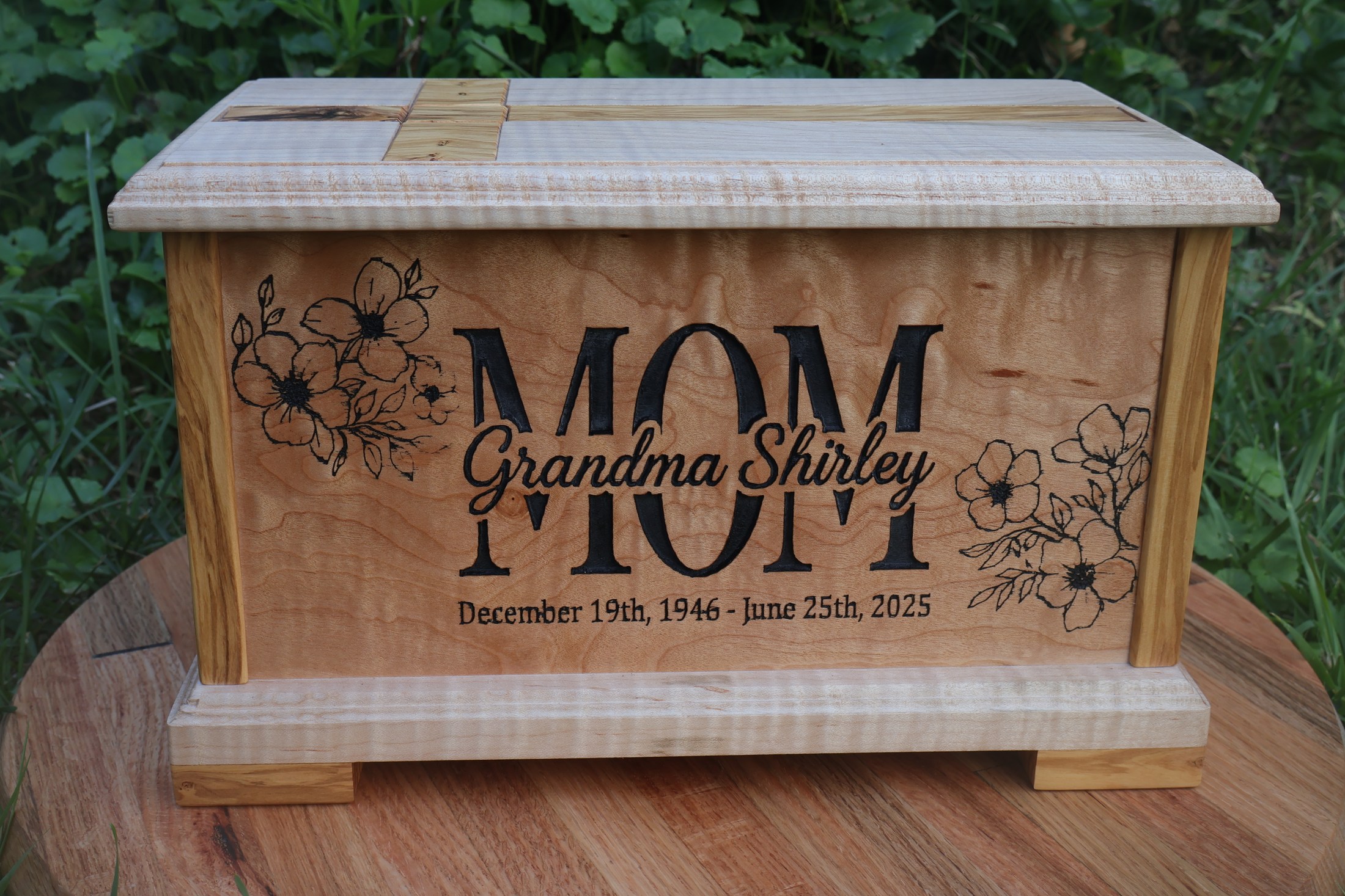 Figured wood memorial box engraved MOM and Grandma Shirley, displayed outdoors on a round wood surface.