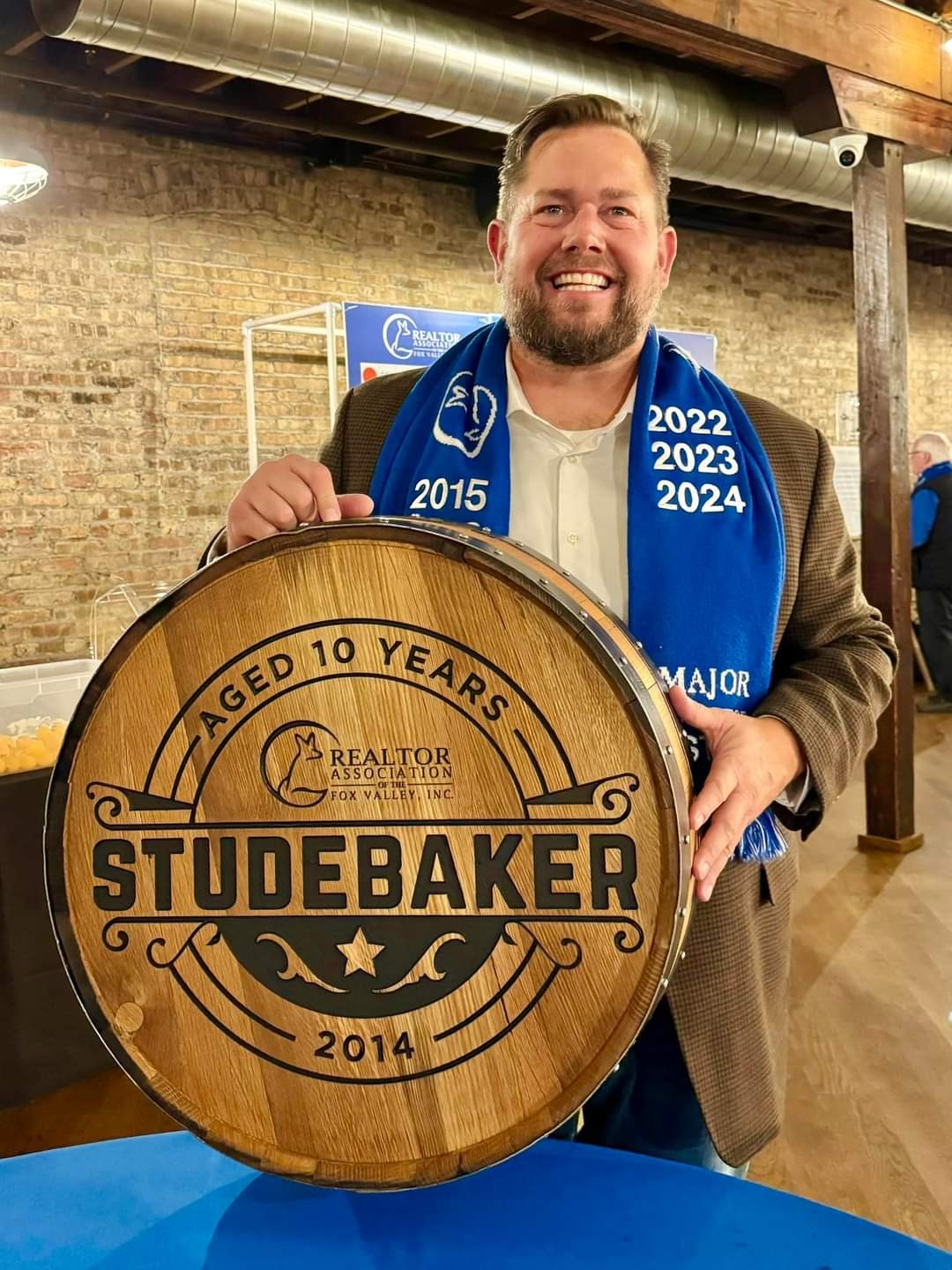 A smiling customer holds an engraved Studebaker barrel cap with an Aged 10 Years message, a Realtor emblem and the year 2014.