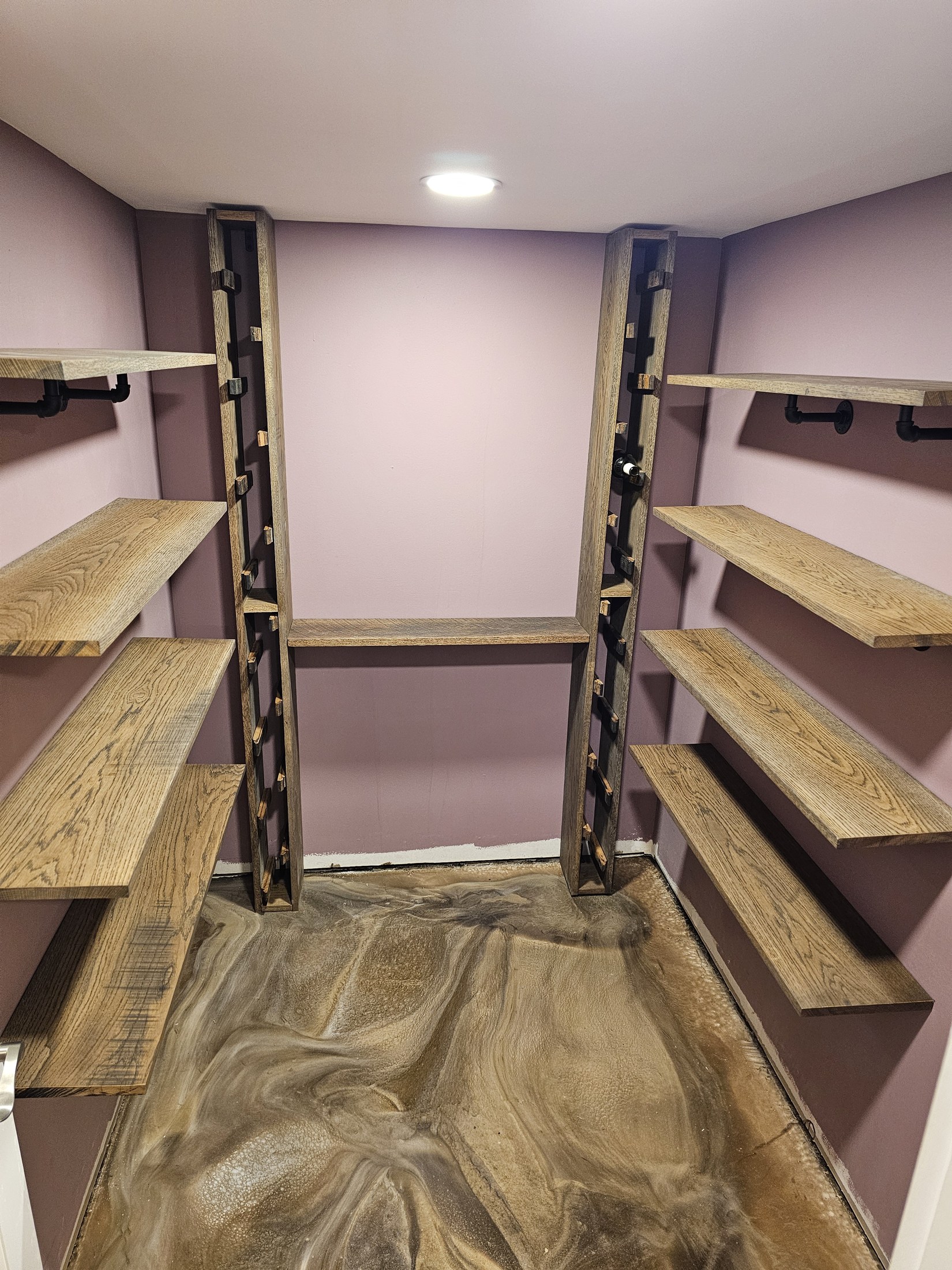 Wood shelves on black pipe brackets line both sides of a narrow room, with two vertical bottle racks against the back wall.